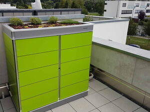 . Ein grüner Schrank mit bepflanztem Dach steht auf einer Terrasse mit Blick auf Wohngebäude und Grünflächen.