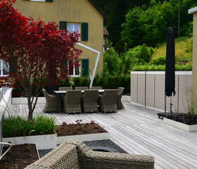 The image depicts an outdoor patio area with a wooden deck. There are wicker chairs and a table set suitable for outdoor dining. A red-leafed tree and some planters are also present on the deck. In the background, there’s a house with a beige exterior and green shutters, as well as greenery and a hill with trees. An umbrella is seen folded near the table.