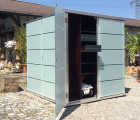. This image shows an outdoor storage structure with its door open. The structure has a modern design with glass panels and appears to have shelves inside. There are various garden elements around it, such as potted plants and decorative objects.