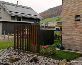 Ein moderner Garten mit Holzzaun, Solarpanelen auf dem Dach, Blumenbeet und grünem Rasen vor einer Holzhütte.