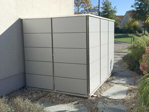 . This image shows a modern, gray outdoor shed, likely made of metal or a similar material. It is situated next to a building and is surrounded by a garden area with stone pathways and some plants. The setting appears to be in a residential area with a street and trees visible in the background.