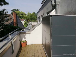 . Eine Terrasse mit Holzboden, Pflanzkübel, Metallgeländer und Häusern im Hintergrund.