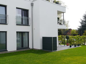 . The image shows a modern residential building with large windows and balconies. The building is white with black window frames. On the ground floor, there is a patio area with potted plants and a small table. The foreground features a well-maintained grassy lawn. There are trees and another building visible in the background.
