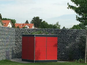 . The image shows a red rectangular structure situated in a garden area. It is near a gray wire mesh fence filled with rocks. The ground is covered with grass, and there are trees and houses with red roofs in the background.