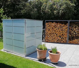 . The image shows a garden scene with a modern storage shed made of frosted glass or similar material on the left. To the right, there is a stack of firewood in a metal frame. In the foreground, there are two potted plants on a tiled patio. The surrounding area is lush with green grass and trees.