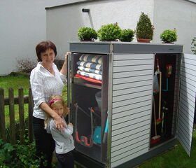 . In the image, there is a woman and a child standing next to an outdoor storage shed. The shed's doors are open, revealing various garden tools inside, such as a broom, rake, and shovel. There are also folded items, possibly garden cushions or mats. On top of the shed, there are a few potted plants. The setting appears to be a garden or backyard area.