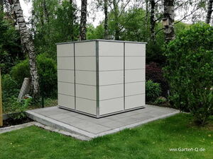 . The image shows a modern, rectangular garden shed or storage unit placed on a paved area in a garden. It has a minimalist design with smooth, light-colored panels and metal edges. The shed is surrounded by grass, trees, and shrubs in a lush green environment.