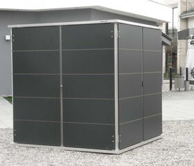 . The image shows a dark-colored metal shed with a modern design. It's located outdoors on a gravel surface, and there are buildings visible in the background. The shed has a sleek appearance with vertical lines and a flat roof.