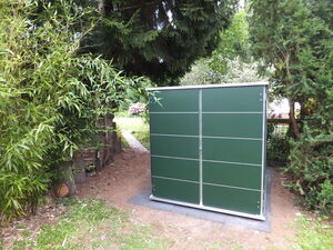 . The image shows a green shed or storage unit located outdoors. It's surrounded by trees and plants, with a pathway leading through the greenery. The area appears to be part of a garden or backyard setting.