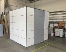 . In the image, there is a large, cube-like structure made of what appears to be white panels with a gray door. It's located in a workshop or industrial setting, with tools and equipment visible in the background. The structure resembles a small room or enclosed area, possibly used for privacy or storage.