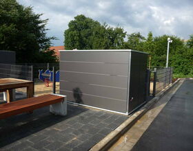 . The image shows an outdoor playground area. There is a gray structure in the center, possibly a storage unit, surrounded by a fence. To the left, there's a wooden bench or table. A children’s play equipment with blue and red colors is visible in the background. There are trees and greenery around the area.