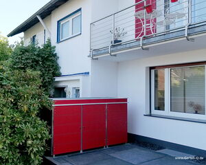 . Ein modernes Haus mit Balkon, rotem Schrank draußen, umgeben von Pflanzen.