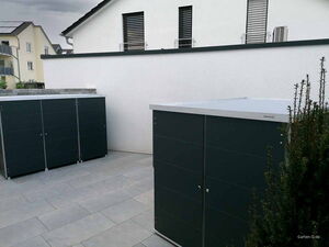 . Zwei moderne dunkle Gartenschränke auf einer Terrasse neben einem weißen Gebäude.