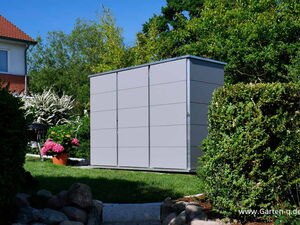 . The image depicts an outdoor garden scene featuring a large, gray storage shed. The shed is situated within a landscaped area, surrounded by well-maintained bushes and flowers. There is a house visible in the background, along with various trees and plants. The setting appears to be a residential garden space.