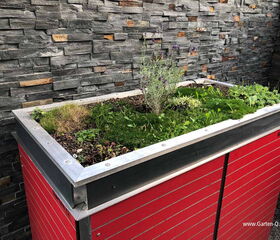 . The image shows a raised garden bed with various plants, including some grass and small shrubs, growing in it. The structure is made of metal with red panels and is placed against a stone wall. The garden bed is designed to be compact and modern, suitable for small spaces or urban gardening.
