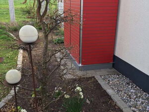 . The image shows a red storage enclosure or box attached to the side of a building. The area around it includes a small garden with a tree and blooming white flowers, possibly daffodils. There are also spherical garden lights on poles. The surroundings have paving and a patch of grassy land.