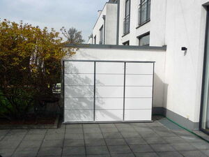 . The image shows a white storage unit or shed on a tiled patio area. The unit is placed against a wall of a building, which appears to be modern with large windows. To the left of the storage unit, there is a hedge or shrubbery in a raised planter.