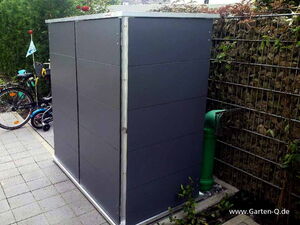 . The image shows a sleek, modern garden storage unit with a rectangular shape, set on a patio. This storage unit has a minimalist design with smooth, dark panels. Next to it on the left are parked bicycles. To the right, there's a wire fence filled with rocks, which resembles a gabion wall. There's also a green pipe with a protective cage near the unit. The ground is paved with square tiles, and there's vegetation visible around the area.