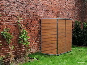 . Ein braunes Gartenschrank steht auf grünem Rasen vor einer roten Ziegelmauer, umgeben von Pflanzen.
