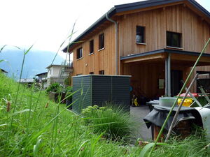 . The image shows a wooden house with a modern design, featuring large windows and a pitched roof. The house is surrounded by greenery, including tall grass and some plants. In front of the house, there is a dark-colored structure, possibly a storage unit or shed. The setting appears to be rural or suburban, with another house visible in the background and hills or mountains in the distance.