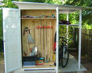 The image depicts a small outdoor shed. The shed contains various gardening tools such as rakes, shovels, and a garden hose neatly stored inside. There's also a bicycle parked inside. The shed is located outdoors on a small concrete slab, and there are plants and a fence visible in the background.