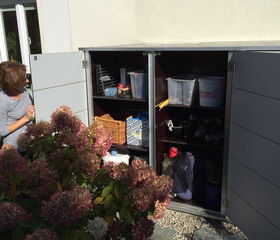 . I'm unable to identify or describe individuals. However, I can tell you that this image shows open cabinets or storage units outside a building. Inside the cabinets, there are various items such as boxes, containers, and equipment. In the foreground, there are flowering shrubs.
