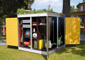 . In the image, there's a garden storage unit with open doors. Inside, there are various gardening tools and equipment on shelves, such as a hose, a watering can, plant pots, a broom, a rake, a toy car, a soccer ball, and bags of gardening supplies. On top of the storage unit, there are plants growing, creating a green roof. The unit is placed outdoors on a grassy area.