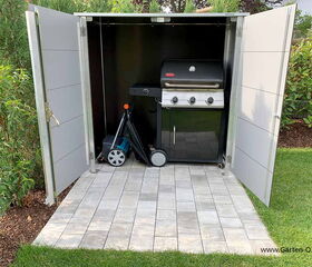 The image shows a storage unit with open doors. Inside, there is a barbecue grill and a lawn mower. The unit is placed on a tiled patio, surrounded by grass and shrubs.