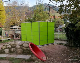 . The image shows an outdoor area with several elements:

1. A green rectangular structure that looks like a garden shed or storage unit.
2. A small playground area with a red slide and rocky climbing surface.
3. Trees with autumn foliage in the background.
4. A fence and some outdoor furniture.
5. Hilly landscape and houses in the distance. 

The setting appears to be a garden or a small park.