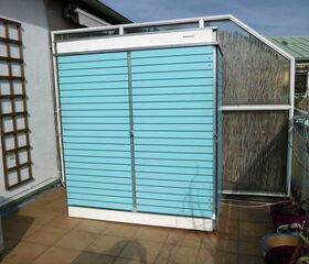 . The image shows a compact, wall-mounted garden storage unit in a light blue color. It is placed on a tiled balcony or patio area. The unit has doors that likely open for storage, and there are some plants and gardening items around it. There's also a wooden trellis on the wall to the left.