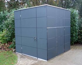 . This image shows a dark gray outdoor structure, possibly a storage shed or utility box, located in a garden or park setting. It has a simple, geometric design with a door and latch on one side. The area around the structure is paved and there are bushes and trees in the background.