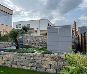 . Ein moderner Garten mit Steinterrassen, Betonstufen, Pflanzen und einem großen grauen Gartenhaus.