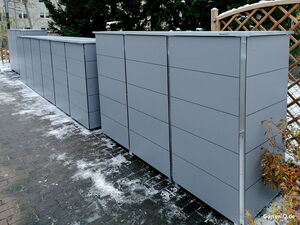 Graue Gartenboxen auf einem schneebedeckten Weg, umgeben von Pflanzen und einem Holzgitter.
