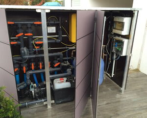 The image shows a technical setup within a cabinet, likely related to water or pool management systems. It includes:

1. Pipes and valves with orange handles for controlling flow.
2. Electrical components and wiring, potentially for monitoring or controlling the system.
3. A container with a liquid, possibly chemicals for water treatment.
4. A pump, suggesting water circulation or filtration.
5. Control boxes mounted on the cabinet door, which might contain electronics for system management.

This setup is typically used for maintaining swimming pools or water features.