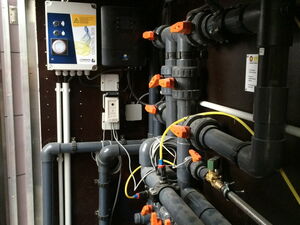 . The image depicts a mechanical room or control area containing a complex array of pipes and valves, likely part of a plumbing or pump system. There is a control panel on the wall with various connections, including electrical and piping components. The orange handles indicate valves for controlling the flow of fluid through the system. Various cables and wires are also visible, suggesting instrumentation or control systems are integrated into the setup.
