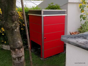 . The image shows a red garden storage unit or box, possibly used for organizing outdoor tools or equipment. It is situated in a garden area, surrounded by greenery and flowers. There is a tree next to the storage unit, and a handle or pole leaning against it.