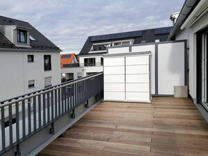 . Eine Dachterrasse mit Holzfußboden, Geländer und Sichtschutz, umgeben von modernen Gebäuden mit Solarpanelen.
