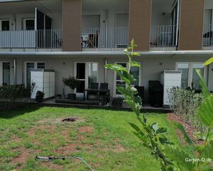 . The image shows a residential area with a two-story building featuring balconies. In the foreground, there's a grassy yard with some bare patches and a garden hose with a sprinkler. The building has outdoor furniture on the balconies and ground floor patios. There are also plants and shrubs lining the outdoor space.