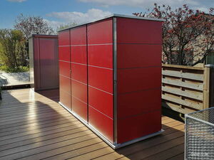 . Ein rotes Outdoor-Metallgerätelager steht auf einer Holzterrasse, umgeben von Holzzaun und Bäumen.