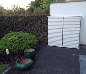 . Ein Garten mit Hecke, zwei leeren Blumentöpfen und einer weißen Wand mit Metalltür.