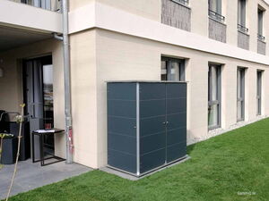 . Ein grauer Gartenschrank steht neben einem Gebäude auf einer Terrasse mit Rasenfläche.