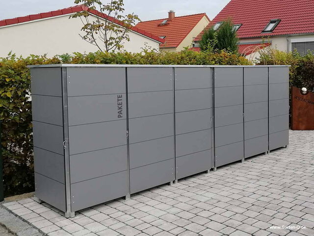 . Eine Reihe grauer Schränke für Pakete steht draußen auf einem gepflasterten Weg vor einigen Häusern.