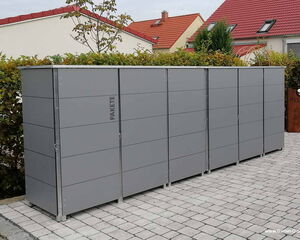 . Mehrere graue Paketboxen auf gepflastertem Boden, mit Hecken und Häusern im Hintergrund.