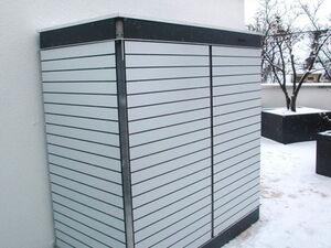 . The image shows an outdoor cabinet or enclosure with horizontal slats, positioned against a building. It appears to be on a snowy surface, suggesting it's winter. There are some planters and a tree visible in the background.