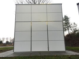The image shows a gray electrical or utility box with a grid panel design on its front. It's in an outdoor setting with grass, trees, and a cloudy sky in the background.