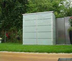 . This image shows a small, modern outdoor storage shed or cabinet situated in a garden. It's made of frosted glass panels with metal framing. Surrounding it are various plants, including some in pots. There's a well-maintained grass lawn and a wooden deck in the foreground. The setting appears contemporary and neatly arranged.