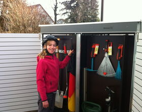 . The image shows a child wearing a helmet and a pink jacket standing in front of a garden storage cabinet. The cabinet contains various gardening tools, such as a rake, a broom, a watering can, and some other hand tools. The interior of the cabinet has hooks and holders for organizing the tools. There is also a flag inside the cabinet.