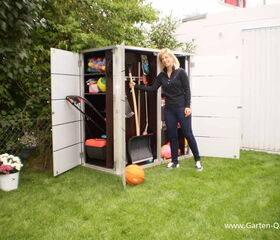 . The image shows a person standing next to an open garden storage unit. Inside the storage, there are various items such as gardening tools, a snow shovel, a rake, some sports equipment like balls, and possibly other outdoor items. There are flowers in pots nearby, and the setting is an outdoor garden or backyard area.