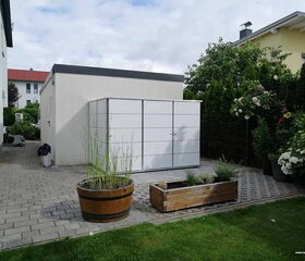 . The image shows an outdoor space with several features:

1. **Buildings**: There appears to be a white structure with a flat roof and another yellow building with a sloped roof in the background.
2. **Metal Structure**: A white, rectangular metal storage unit or cabinets.
3. **Planters**: There are two planters, one round wooden barrel and one rectangular wooden box, containing various plants.
4. **Pathway**: Paved with rectangular stones.
5. **Garden**: Includes various plants and bushes, some with flowers.
6. **Lawn**: A small grass area is visible in the foreground.

The setting looks like a residential backyard or garden area.