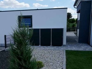 . The image shows a modern, minimalistic garden with some elements. Here's what I can describe:

1. A small landscaped area with light-colored gravel and a trimmed shrub.
2. Neatly arranged grassy lawns on either side of the gravel path.
3. A building with white walls and a flat roof, with what seems to be storage units or doors attached to it.
4. Clear blue sky with scattered clouds.

The setting is clean and organized, typical of contemporary outdoor design.